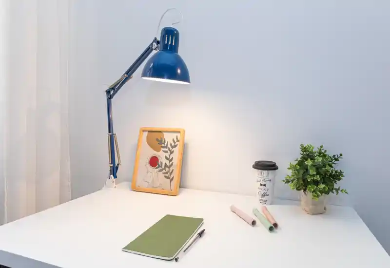 Angolo scrivania luminoso e minimale con lampada blu regolabile, quadro incorniciato, pianta in vaso e cancelleria — perfetto per mostrare un piccolo spazio di lavoro.