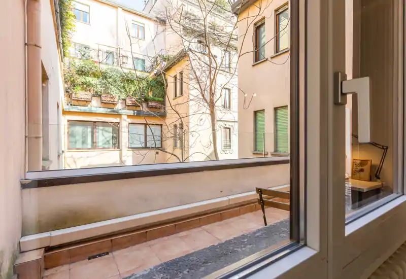 Piccolo balcone privato che affaccia su un tranquillo cortile interno; pavimento in piastrelle, parapetto basso e seduta visibili attraverso la finestra aperta — spazio esterno luminoso e riparato.