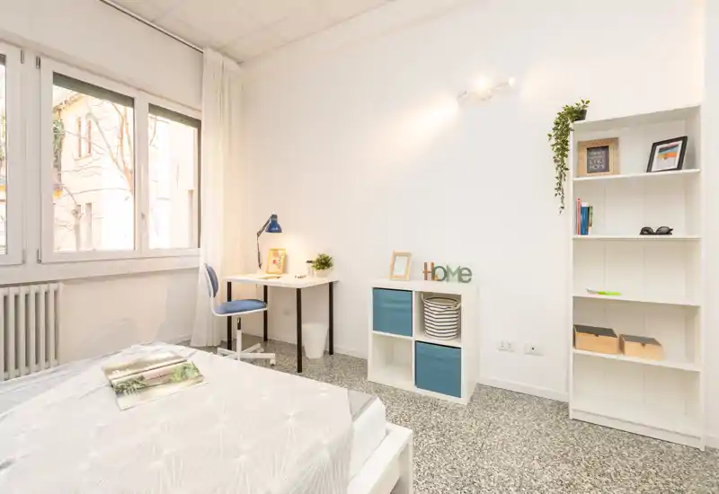 Camera singola luminosa e moderna con letto ben rifatto, piccolo scrittoio vicino alla finestra e scaffalatura ordinata — ideale per uno studente o professionista.