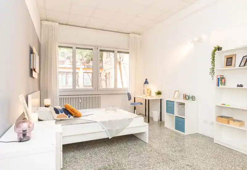 Camera da letto luminosa e minimal con un grande letto, scrivania vicino alla finestra, scaffalatura e cuscini decorativi: ideale per una persona o una coppia.