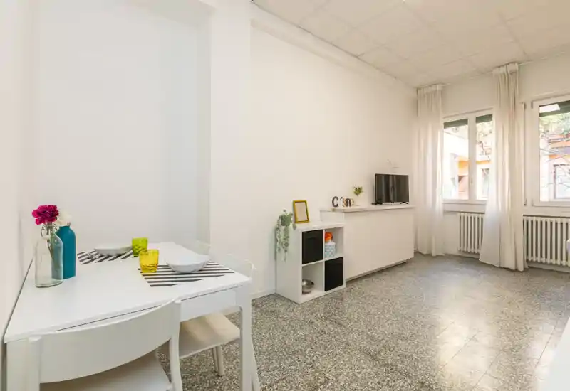 Zona giorno luminosa e minimal con un piccolo tavolo da pranzo, mobile TV e grandi finestre che lasciano entrare luce naturale; arredi neutri e pavimento in graniglia donano un senso di pulizia e ampiezza.