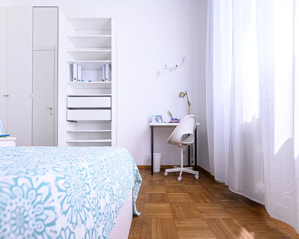 Bright, minimal bedroom with a patterned turquoise bedspread, built-in shelving and a small study desk by a large window that fills the room with natural light.