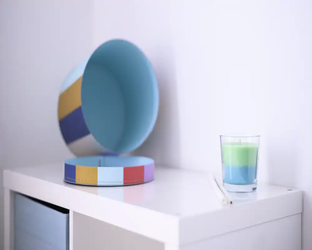 Close-up of a tidy surface with a colorful round box and a layered green-blue candle on a white cabinet — a small decorative detail suitable for a living area or bedroom accent.