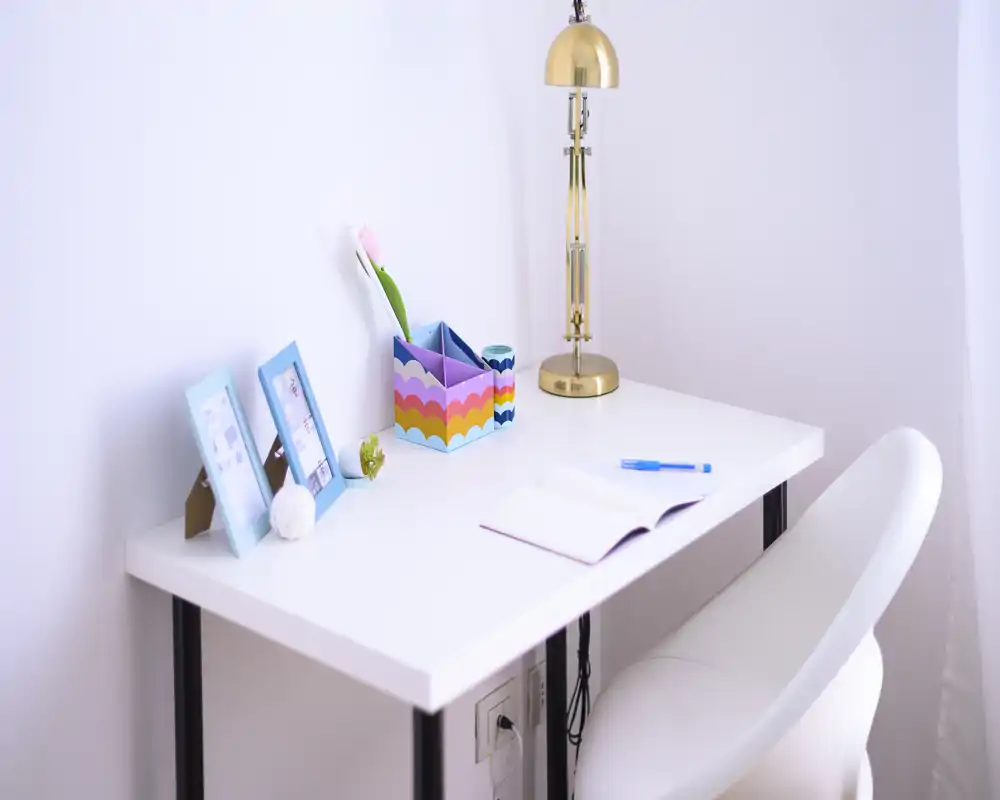 Bright, minimalist desk nook with a white table, comfortable chair, brass lamp and small decorative items — ideal for focused work or study.