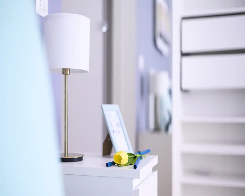 Close-up of a bedroom bedside table with a lamp, picture frame and a decorative paper flower; soft, bright tones create a calm atmosphere.