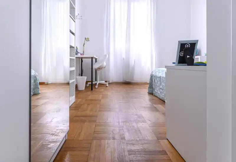 Bright, minimal bedroom with parquet flooring, twin beds partially visible, a small desk and large windows letting in soft natural light.