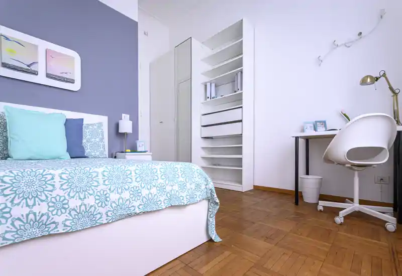 Bright, modern bedroom with a patterned turquoise bedspread, layered pillows and a compact workspace with desk and chair—stylish and well-lit sleeping area.