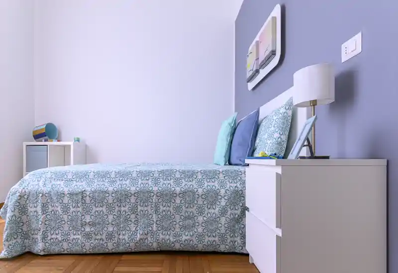 Bright, stylish bedroom with a patterned teal bedspread, layered pillows and a white bedside table with lamp. Clean lines, soft colors and wooden floor give a calm, inviting atmosphere ideal for rental photos.