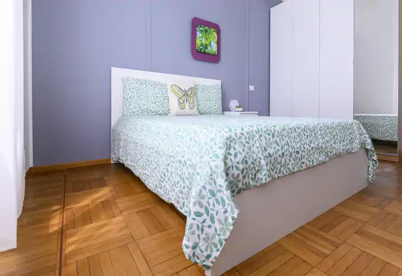 Bright, neatly styled bedroom with a queen bed dressed in a green leafy-pattern duvet, matching pillows and a simple white headboard. Natural light fills the room, highlighting the parquet floor and a mirrored wardrobe that adds depth.