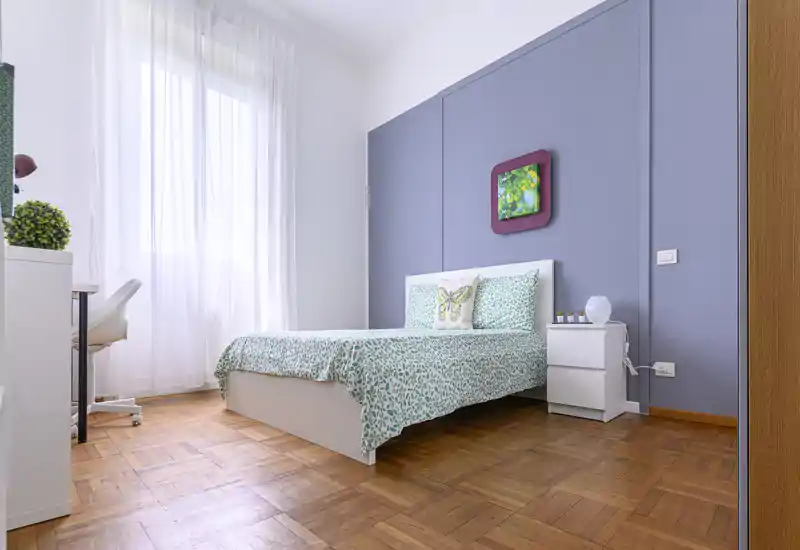 Bright, neatly staged bedroom with a double bed, patterned bedding and a bedside table; large window provides abundant natural light.