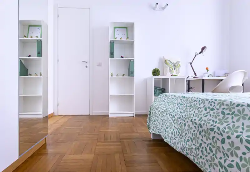 Bright, tidy bedroom with a patterned green-and-white bedspread, desk and shelving — minimal, modern styling and warm parquet flooring.