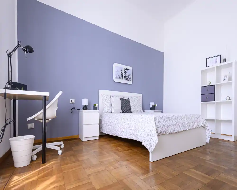 Bright and tidy bedroom with a double bed, bedside tables, a small workspace and shelving; neutral décor and wooden parquet floor create a calm, welcoming atmosphere.