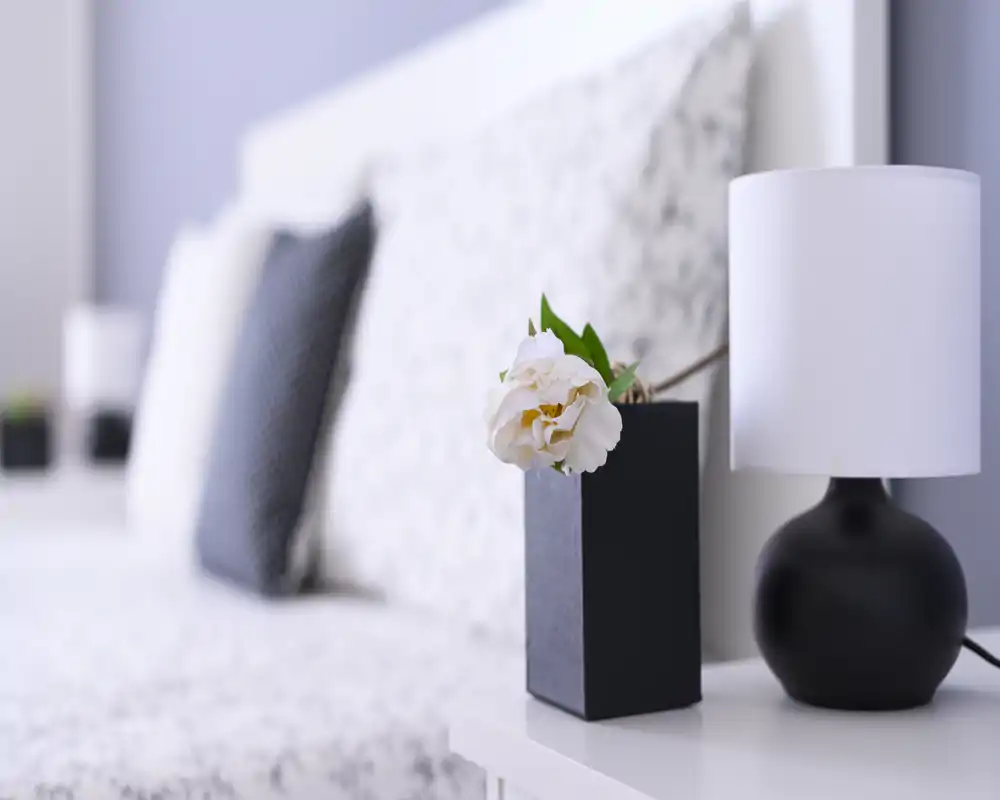 Bright, styled bedroom close-up showing a bedside table with a lamp and flower vase; soft-focus headboard and pillows create a calm, inviting atmosphere.