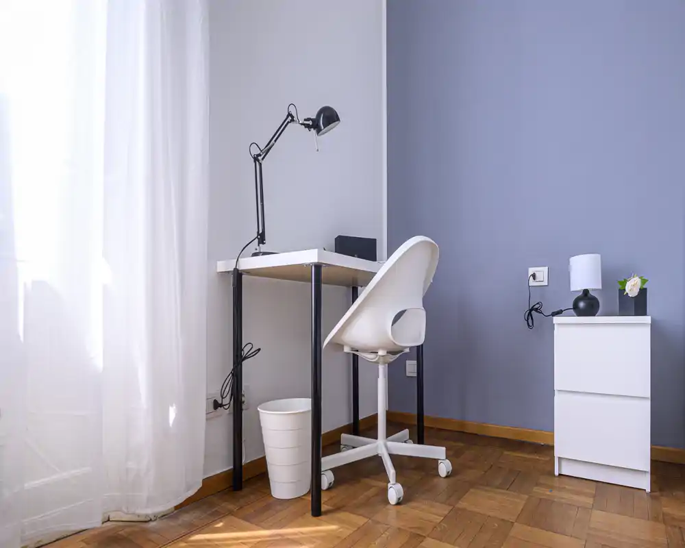 Bright, minimal corner workspace with a white desk, ergonomic chair and a modern lamp next to a sheer-curtained window; clean parquet flooring and a small dresser with a lamp and plant complete the airy living area.
