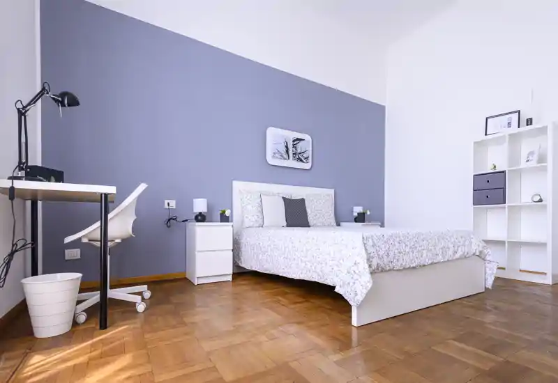 Bright and tidy bedroom with a double bed, bedside tables, a small workspace and shelving; neutral décor and wooden parquet floor create a calm, welcoming atmosphere.