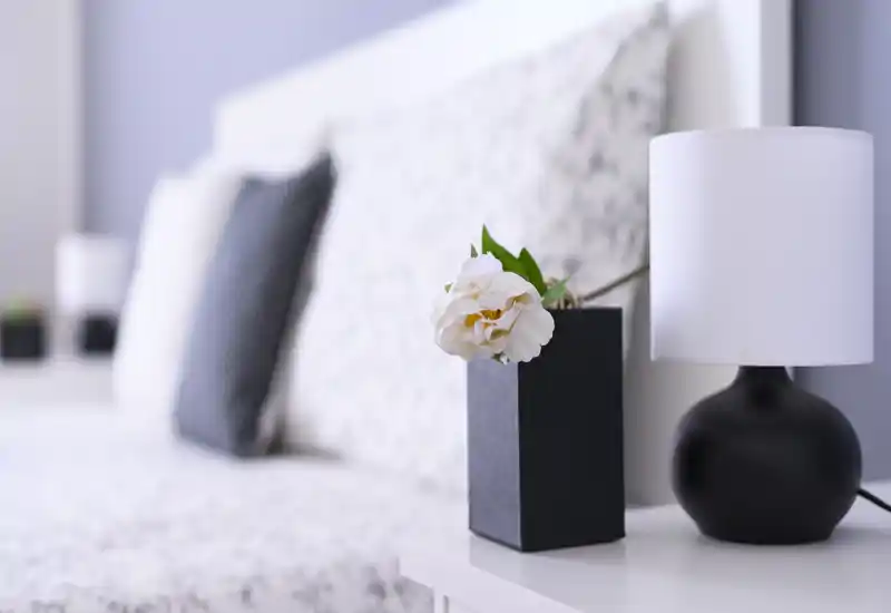 Bright, styled bedroom close-up showing a bedside table with a lamp and flower vase; soft-focus headboard and pillows create a calm, inviting atmosphere.