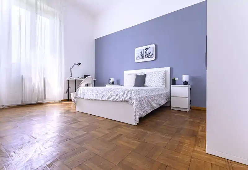 Bright, modern bedroom with a large bed centered against a soft blue accent wall, parquet flooring and a workspace by the window — ideal for rental photos highlighting space and natural light.