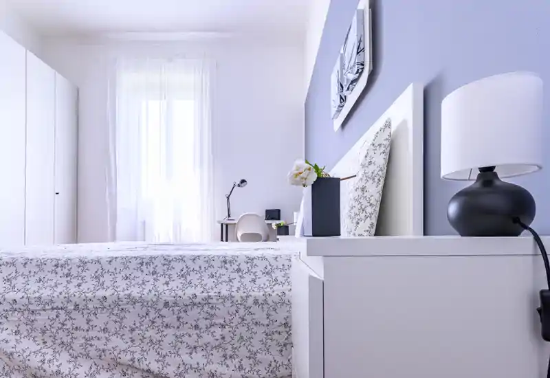 Bright, modern bedroom with a patterned double bed, bedside table with lamp and decorative flowers, and a small workspace by a sheer-curtained window.