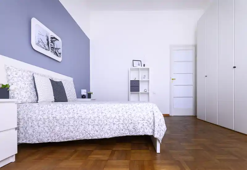 Bright, modern bedroom with a large bed dressed in patterned linens, coordinated cushions and minimalist shelving; wooden parquet flooring and built-in wardrobes create a neat, airy space.