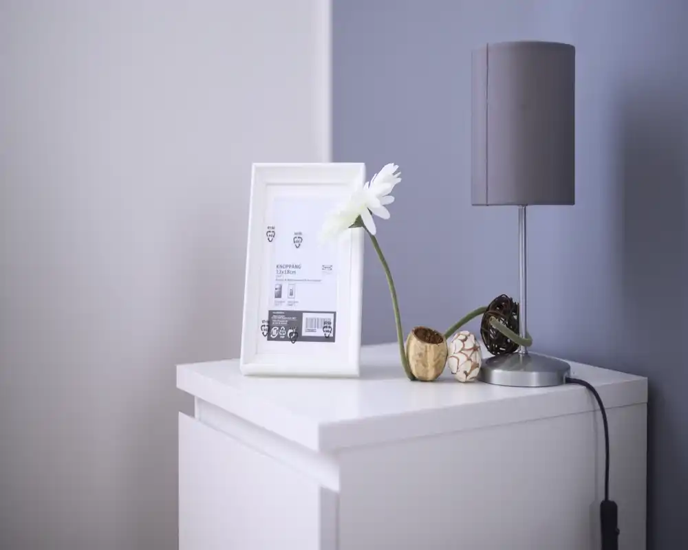 Detalle de mesita de noche minimalista con una lámpara, objetos decorativos y un marco sobre una pared de tonos suaves: un acento moderno y tranquilo del dormitorio.
