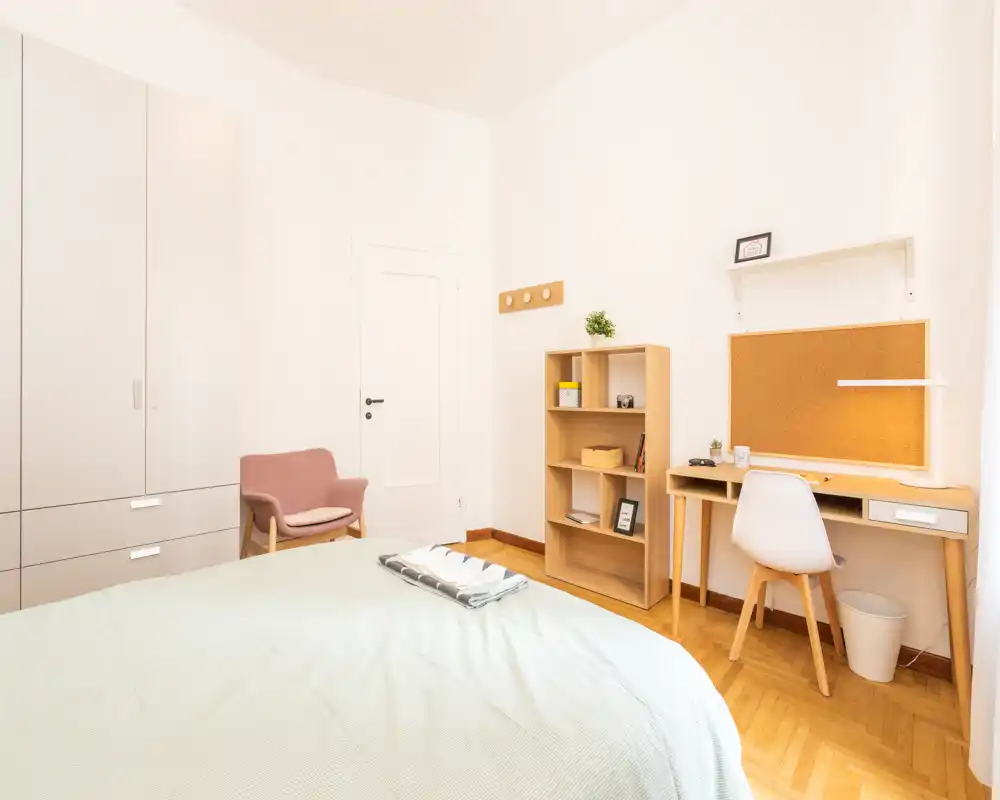 Camera da letto luminosa e minimal con un grande letto in primo piano, scrivania e scaffale, pavimento in legno chiaro: ben illuminata e accogliente per annunci.