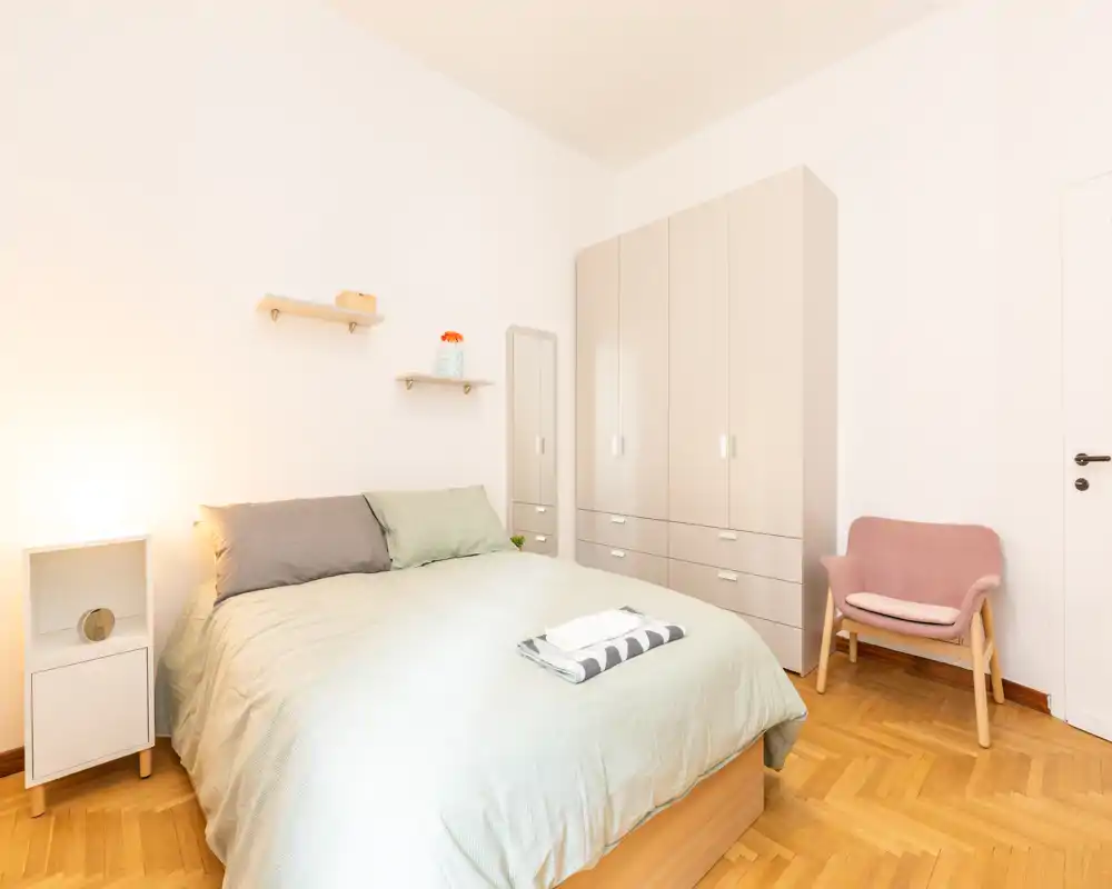 Camera da letto luminosa e ben arredata con letto matrimoniale rifatto, armadio capiente, specchio a figura intera e una comoda poltroncina — perfetta per annunci di affitto.