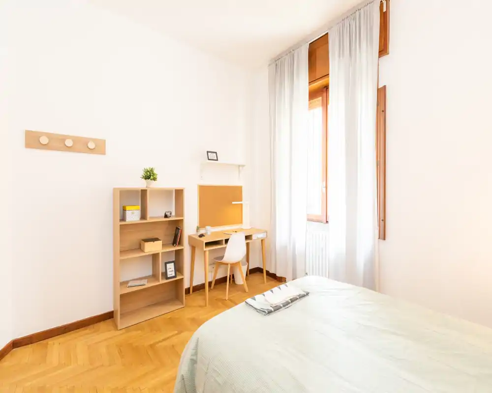 Camera da letto luminosa e minimale con parquet in legno, letto ben rifatto e una scrivania vicino alla finestra — ideale per una persona.
