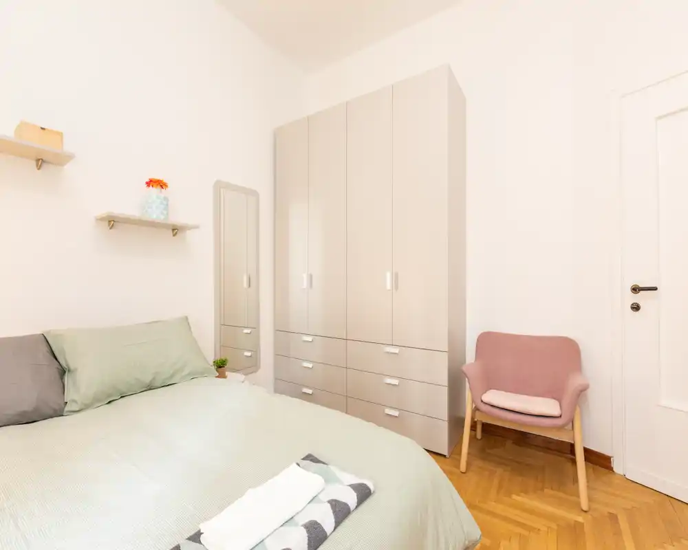 Camera da letto luminosa e ben allestita con un grande letto, armadio alto e una poltroncina accogliente; palette neutra e arredo minimale donano un ambiente tranquillo.