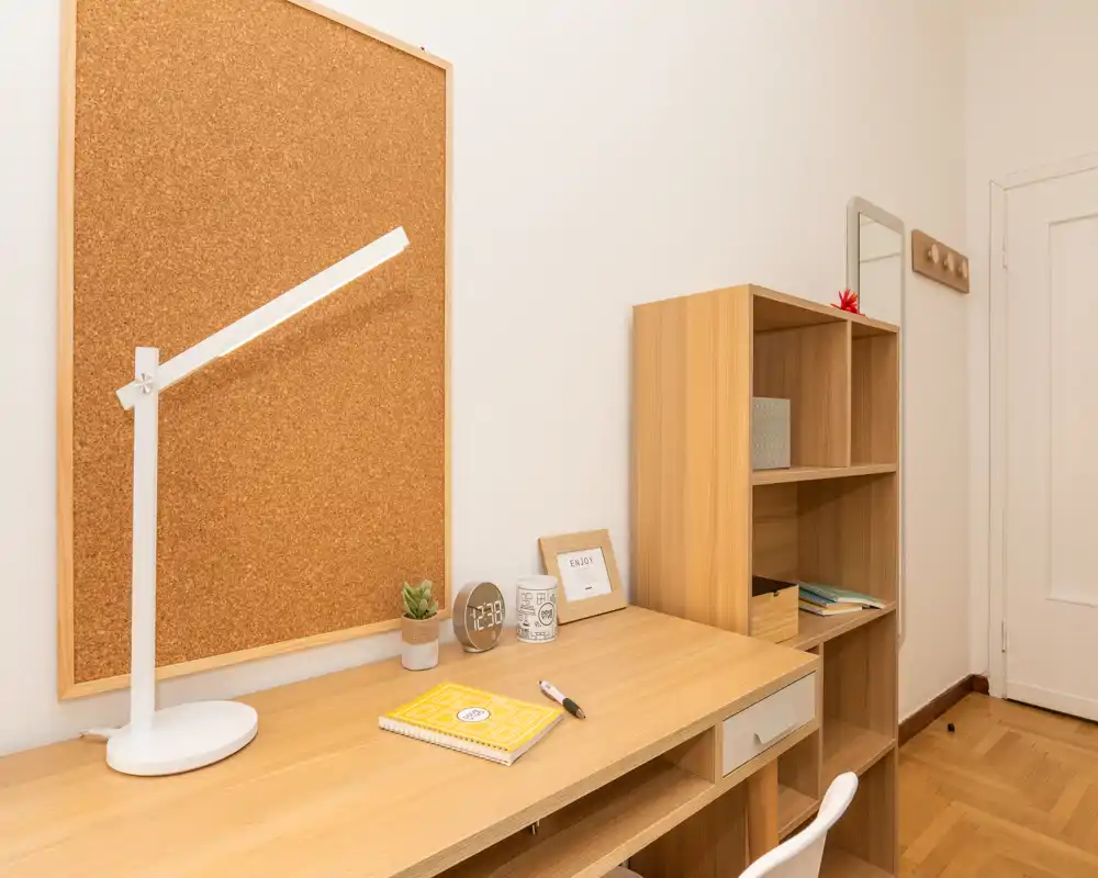 Compact, well-lit study corner with a wooden desk, cork noticeboard, shelving unit and a white task chair — ideal for focused work or studying.