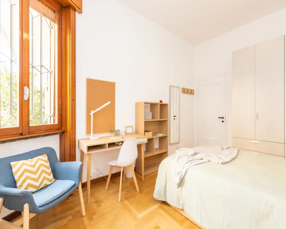 Bright, neatly styled bedroom with a double bed, natural wood furniture and a cozy armchair by a large window allowing abundant daylight.