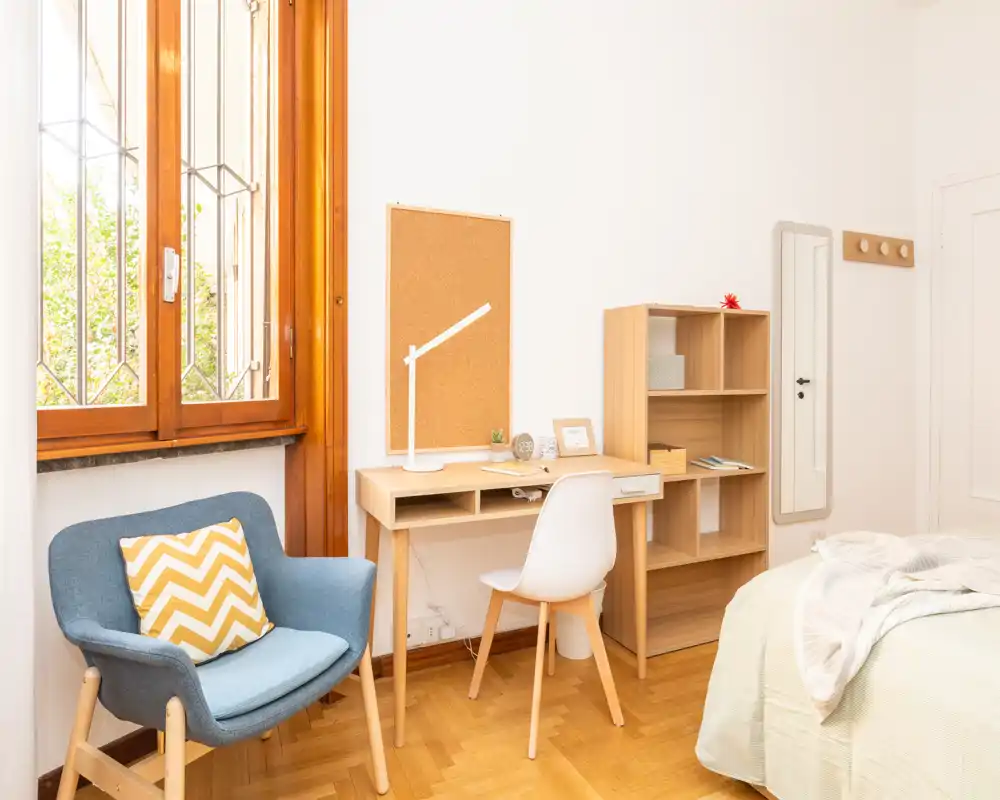 Sunny, neatly staged bedroom corner with a visible bed edge, a small wooden desk and shelving, and a cozy armchair by a large window—ideal for a work-and-relax setup.