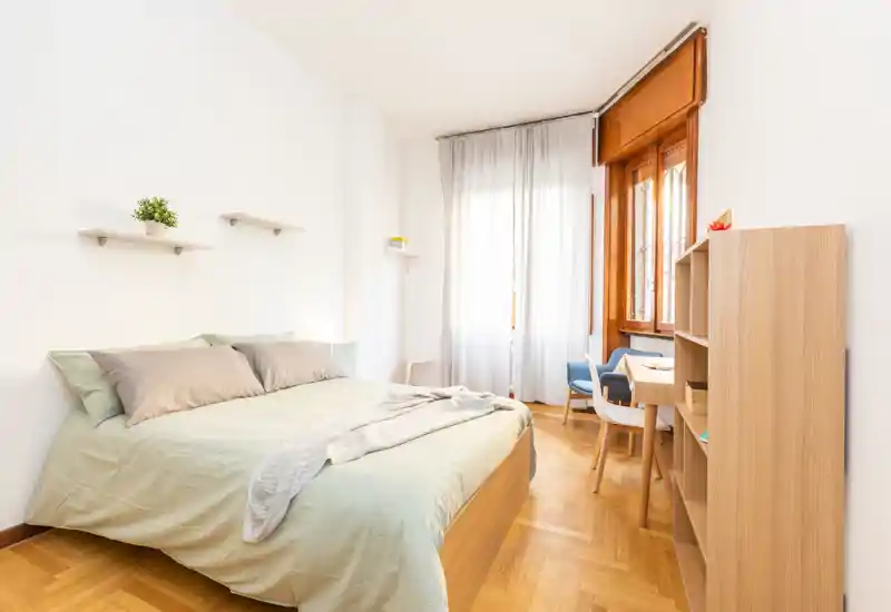 Dormitorio luminoso y bien decorado con cama doble, ropa de cama suave y luz natural de una gran ventana. Estantes sencillos y un área de escritorio lo hacen funcional para vivir y trabajar.