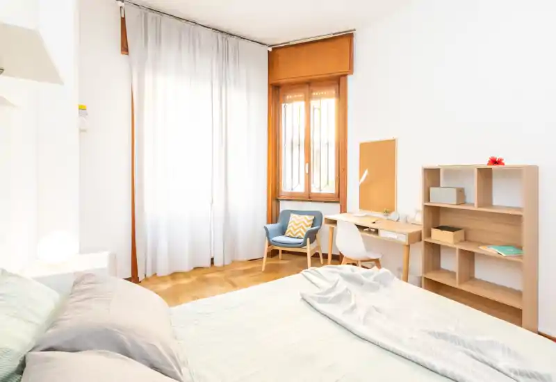 Dormitorio luminoso y bien decorado con una cama grande, ropa de cama suave y un rincón de lectura junto a la ventana. La luz natural, el suelo de madera y los muebles sencillos crean un ambiente acogedor perfecto para anuncios.