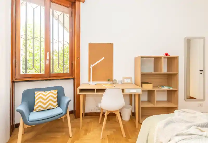 Rincón de dormitorio luminoso y aireado con escritorio pequeño, estantería y un sillón cómodo junto a una gran ventana; ropa de cama parcialmente visible que aporta un ambiente acogedor y funcional para descansar y trabajar.