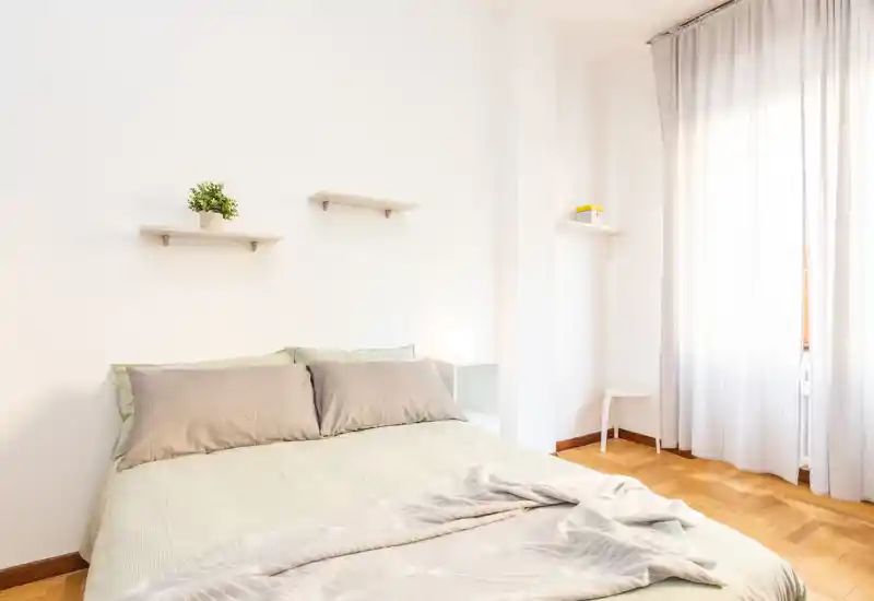 Dormitorio luminoso y minimalista con una cama doble bien hecha, almohadas suaves y luz natural de una gran ventana que crea un ambiente tranquilo y acogedor.