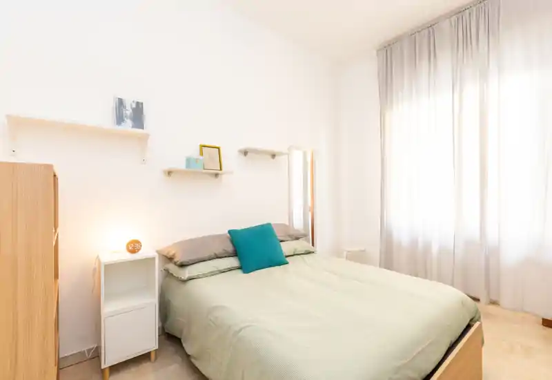 Bright, minimalist bedroom with a neatly made double bed, soft linen, a teal accent cushion and natural light from a large curtained window.