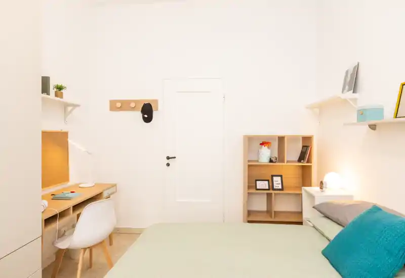 Bright, minimal bedroom with a neatly made bed, bedside lamp, shelving and a small desk — ideal for a single occupant or student.