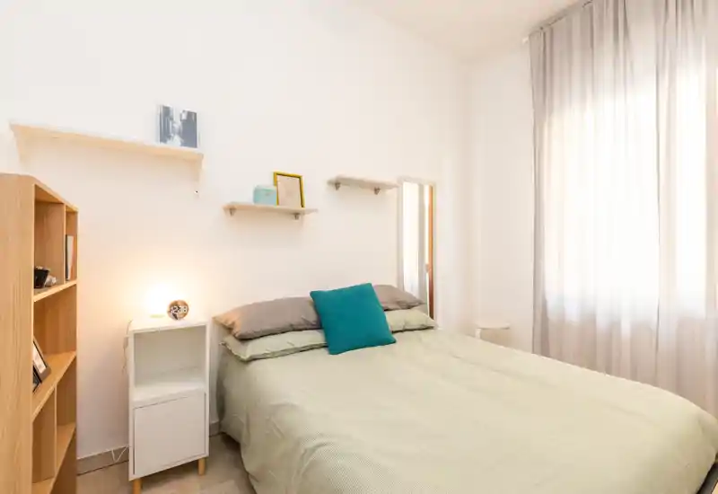 Bright, tidy bedroom with a double bed, soft green bedding, decorative cushion and natural light from a large window; simple shelving and a bedside table complete the cozy layout.