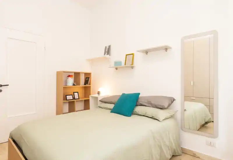 Bright, neatly staged bedroom with a double bed, soft linens and a teal accent pillow. Minimal shelving and a full-length mirror create a clean, welcoming feel — ideal for rental listings.