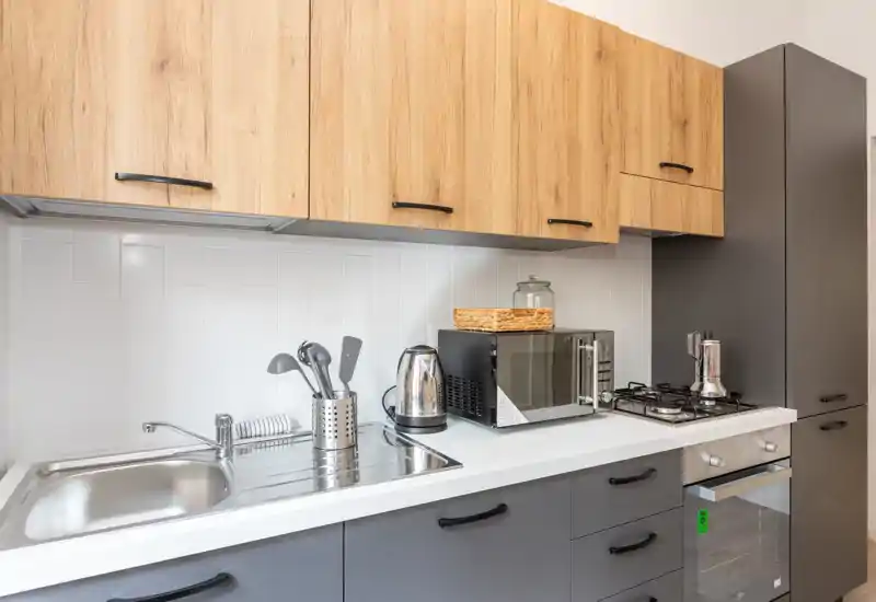 Cocina compacta y moderna con armarios superiores efecto madera, muebles inferiores oscuros, fregadero de acero, placa de gas y electrodomésticos de encimera; luminosa y lista para usar.