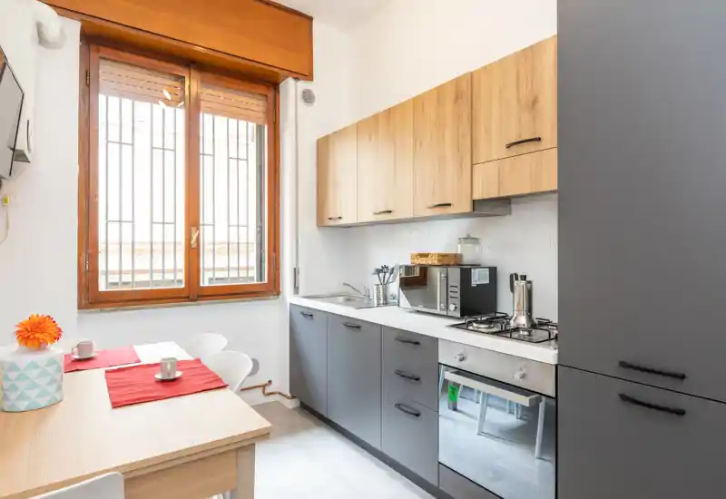 Cocina moderna y luminosa con muebles bajos gris mate, armarios superiores de madera, placa de gas y horno integrado. Una mesa pequeña junto a la ventana forma un acogedor rincón para desayunar.