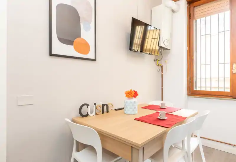 Rincón comedor luminoso y bien presentado con mesa de madera para dos, jarrón decorativo y cuadro moderno — ideal para anuncios de apartamentos pequeños.