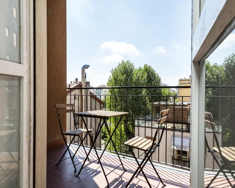 Balcon ensoleillé avec un petit ensemble bistrot en bois et métal, garde-corps en fer et vue sur les arbres et les toits voisins — idéal pour le café du matin.