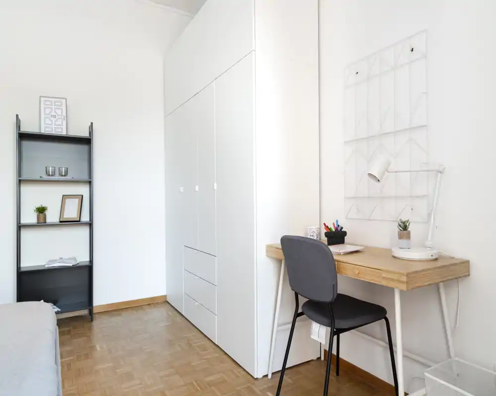 Coin chambre minimaliste et lumineux avec une armoire blanche, un petit bureau en bois et une étagère simple ; une partie du lit est visible à gauche, convenant pour une photo d'annonce courte durée.