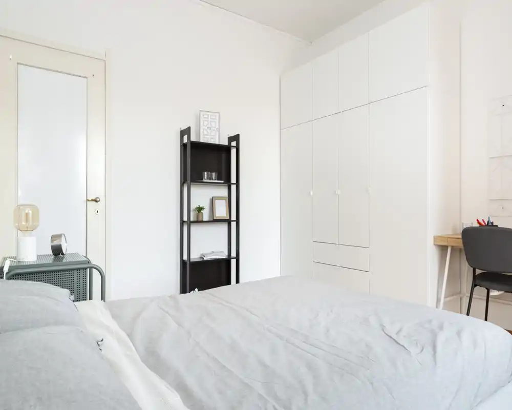 Chambre moderne et lumineuse avec un grand lit au premier plan, une armoire blanche encastrée et un petit bureau. Tons neutres et décoration sobre pour une atmosphère calme et accueillante.