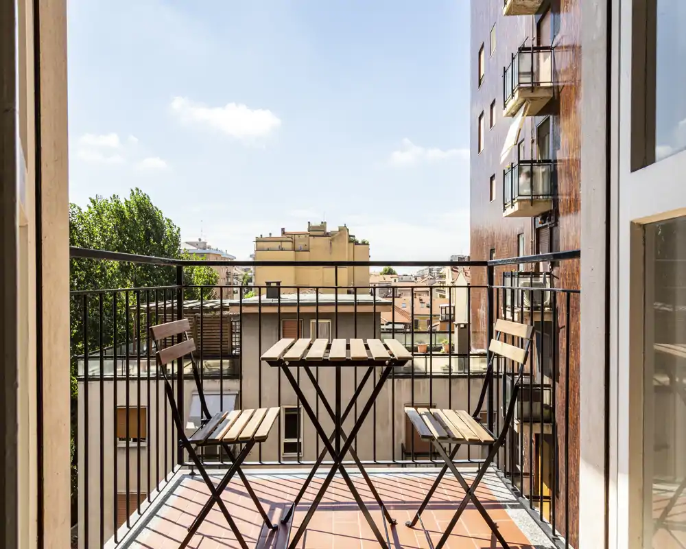 Balcón soleado en la ciudad con pequeña mesa bistró de madera y dos sillas, suelo de baldosa y barandilla de metal — ideal para un café matutino con vistas urbanas.