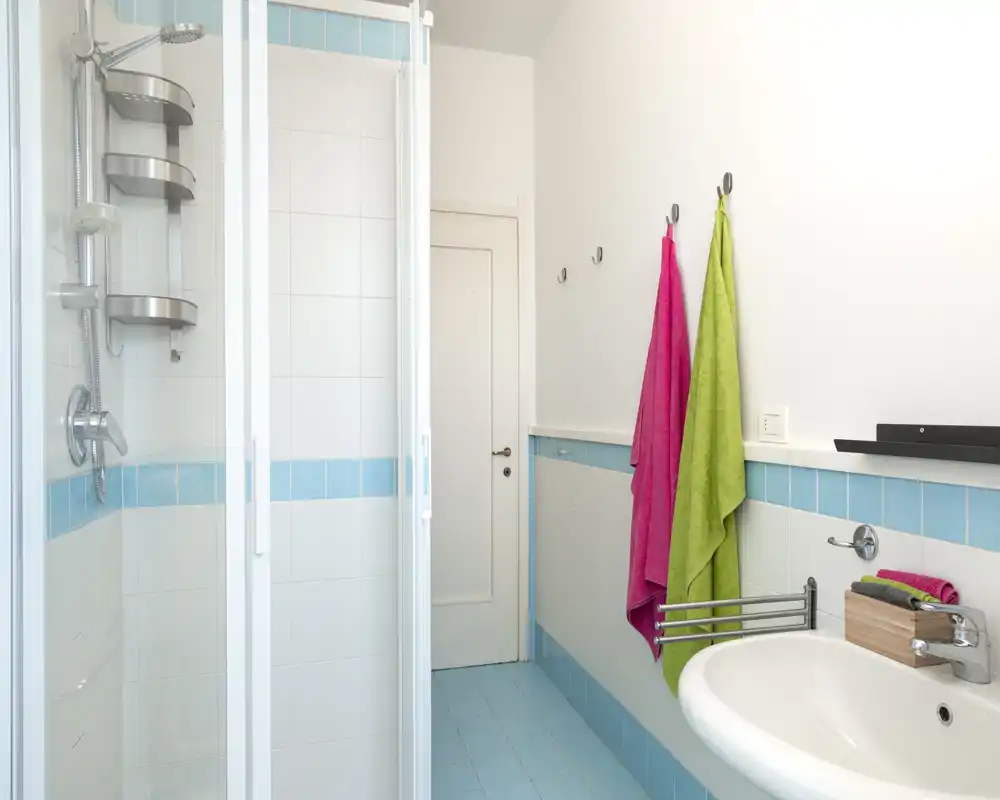 Salle de bains lumineuse et moderne avec cabine de douche carrelée, lavabo mural et serviettes colorées; carrelage blanc et bleu clair pour une ambiance fraîche.