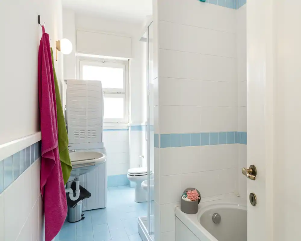 Petite salle de bains lumineuse avec carrelage bleu et blanc, baignoire, cabine de douche, lavabo et WC ; des serviettes colorées ajoutent une touche fraîche.