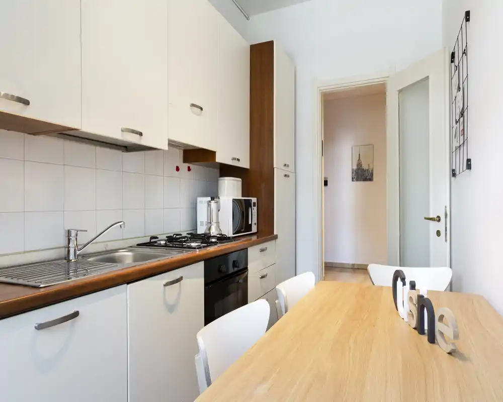 Cuisine moderne et lumineuse avec plan de travail en bois et placards blancs, équipée d'un évier, d'une plaque à gaz, d'un four et d'un micro-ondes. Une table en bois avec des chaises blanches forme un coin repas convivial.