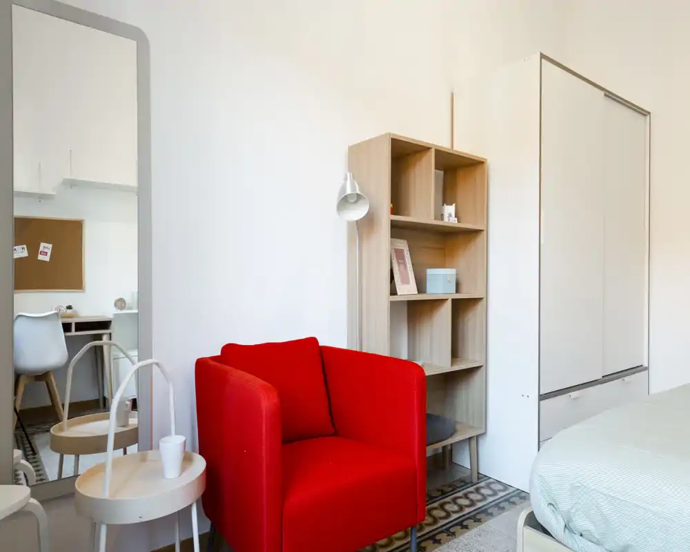 Bright, modern bedroom corner with a vivid red armchair, open shelving and a partially visible bed. Neutral walls and patterned floor tile give a clean, stylish look.